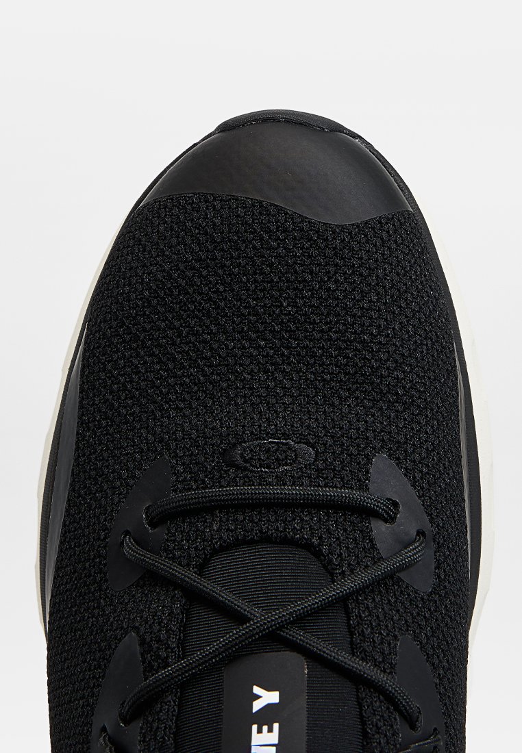 Zapatilla deportiva negra con parte superior de tela texturizada, puntera negra y suave, y suela blanca en contraste. Presenta cordones negros y detalles del logo.