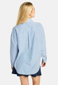 Mėlyna ir balta dryžuota marškinėliai su sagomis, ilgomis rankovėmis, laisvo silueto, su nugaros klostėmis ir apvalinta apačia. Dėvimi ant tamsiai mėlynos spalvos sijono.