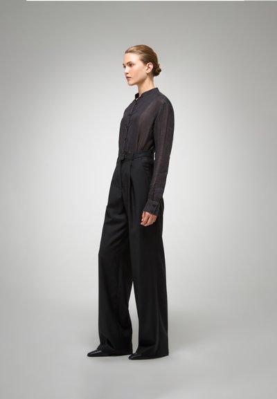Zwarte, doorschijnende button-up shirt met lange mouwen en hoge kraag, gecombineerd met hoge taille, wijde zwarte broek en puntige zwarte schoenen.