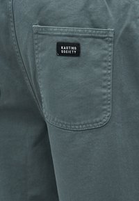 Close-up of gray fabric pants featuring a rectangular back pocket with a black label reading "KAOTIKO SOCIETY" sewn above the pocket.