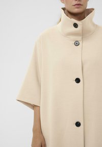 Beige oversized frakke med høj krave, korte ærmer og tre sorte knapper foran. Glat, struktureret stof.