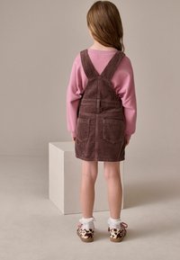 Brun corduroy overallkjole med to forlommer, brugt over en lyserød langærmet skjorte. Brun og hvidprikkede sko med snørebåndssokker.
