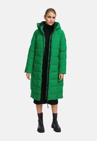 Abrigo largo acolchado verde con capucha, que presenta un cierre frontal, detalles en negro y un diseño acolchado. Lleva puesto sobre un vestido de punto negro y botas.