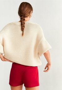 Chunky pletený krémový svetr s krátkým rukávem, spárovaný s červenými šortkami s dvěma zadními kapsami, pohled zezadu. Viditelný texturovaný design.
