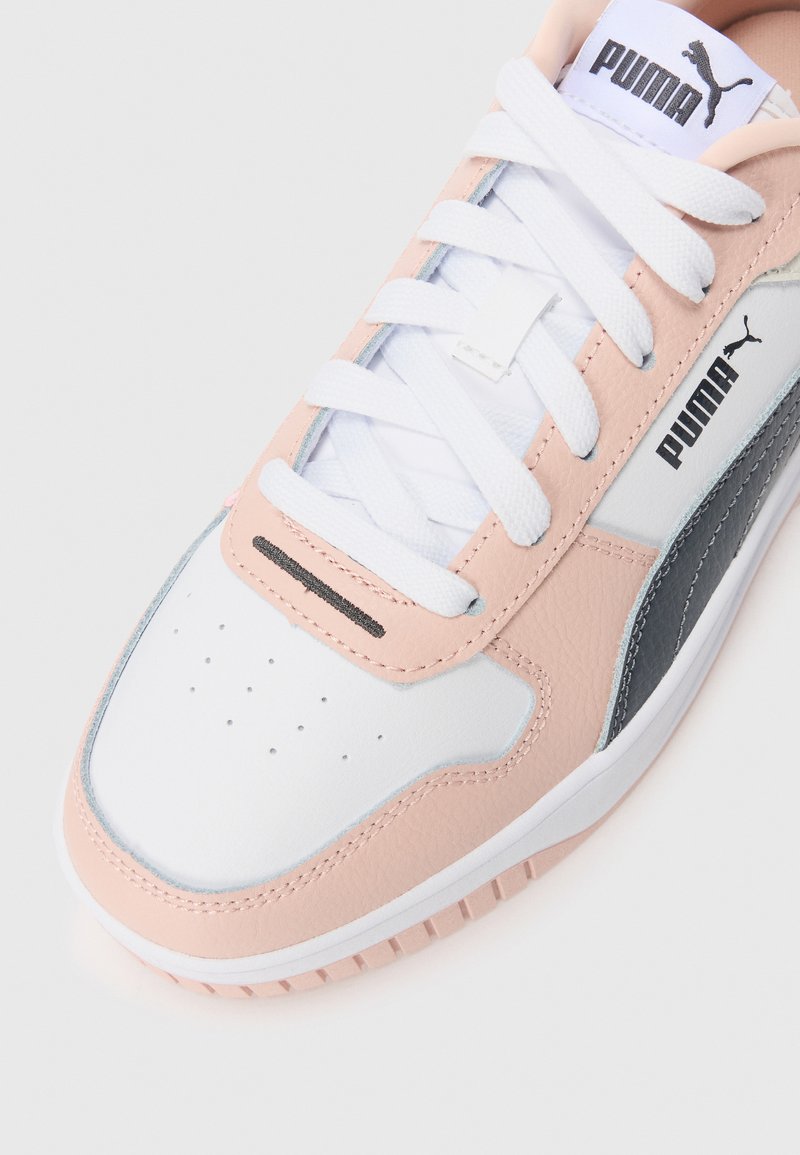 Zapatilla Puma en blanco y rosa claro con cordones blancos, puntera perforada y logo negro de Puma en el lateral y la lengüeta, aislada sobre un fondo blanco.