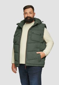 Grüne gepolsterte Weste mit Kapuze, ausgestattet mit zwei Brusttaschen und einem Reißverschluss auf der Vorderseite. Darüber ein leichter Pullover getragen, kombiniert mit blauen Jeans.