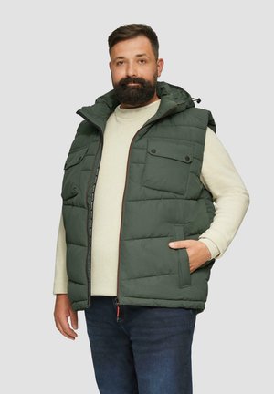 Groene gewatteerde vest met capuchon, voorzien van twee borstzakken en een ritssluiting aan de voorkant. Draagt over een lichte trui, gecombineerd met een blauwe spijkerbroek.