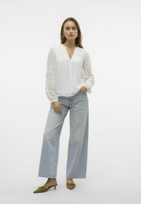 Blusa bianca con dettagli in pizzo su maniche, abbinata a jeans a gamba larga blu chiaro. Il modello indossa ballerine verde olive a punta.