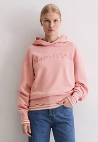 Kobieta w różowym hoodie Marc O'Polo na paskowanej koszuli i niebieskich dżinsach, stoi przy jasnej, teksturowanej ścianie.
