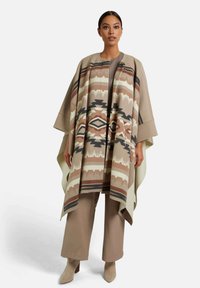 Beige gemustertes Poncho mit geometrischen Designs in Braun und Schwarz, mit offener Front und weiten Ärmeln, getragen über einem einfarbigen Outfit.