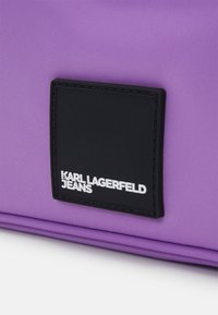 Etiqueta cuadrada negra con texto blanco "KARL LAGERFELD JEANS" sobre fondo de tela morada.