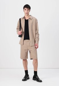 Beige Knopfhemd mit einer Brusttasche, kombiniert mit beigen gestreiften Shorts. Das Modell trägt ein schwarzes T-Shirt und schwarze Schuhe mit Socken.