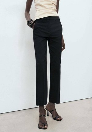 Person, die schwarze Hose mit geradem Bein, beiges Oberteil, schwarze Sandalen mit Absatz und schwarze Armbänder trägt, steht an einer weißen Wand.