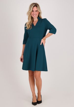 Lachende vrouw in een tealkleurige knielange jurk met driekwart mouwen en zwarte hakken, staand met één hand op haar heup.