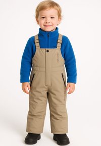 Tan sneeuwoverall met verstelbare bandjes, met rits zakken en logo-detail, gecombineerd met een blauwe fleecesweater. Zwarte laarzen maken de outfit compleet.