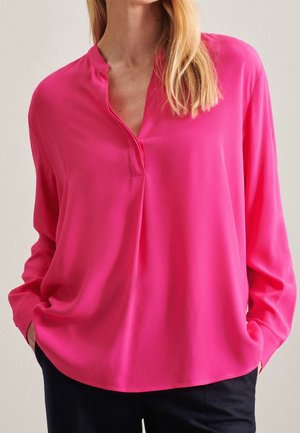 Kvinde iført en knaldrød, langærmet bluse med V-formet halsudskæring og sorte bukser, hænderne i lommerne, foran en ensfarvet baggrund.