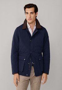 Marineblaue gesteppte Jacke mit braunem cordstoffartigen Kragen, Reißverschluss vorne und Taschen mit Druckknopfverschluss. Strukturiertes Material mit taillierter Passform.