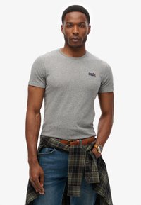 Grå kortærmet t-shirt med rund hals, der har et lille logo i marineblå. Båret sammen med blå denim jeans og en bundet ternet skjorte om livet.