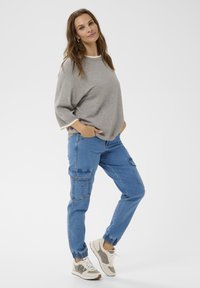 Ženska z dolgimi rjavimi lasmi, oblečena v sivo ohlapno jopico, modre cargo hlače in bež tene, stoji s rokami v žepih na belem ozadju.