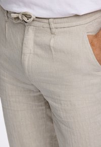 Lichtbeige broek van gestructureerde stof met een visgraatpatroon. Voorzien van een verstelbare koordtaille en standaard zijzakken.
