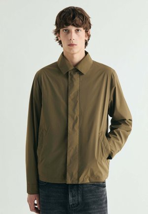 Joven con cabello corto y castaño, vestido con una chaqueta verde oliva con botones delanteros ocultos y vaqueros oscuros, de pie frente a un fondo liso.