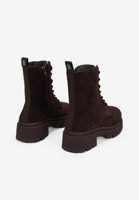 Botas de tobillo de ante marrón con una suela de goma gruesa, cierre de cordones en la parte delantera y cremallera lateral. Paneles ribeteados verticales y logotipo en el talón.