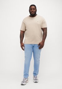 Beige kortermet t-skjorte med rund hals, kombinert med lyseblå jeans og grå sneakers. Merkbart tatoveringer på armene er synlige.