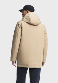 Beige gewatteerde jas met een hoge kraag en een geïntegreerde capuchon, met een rechte snit en een label op de linker schouder.