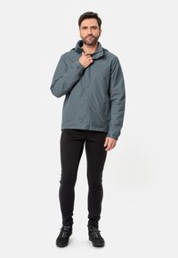 Giacca impermeabile grigia con cappuccio, polsini elastici e zip frontale. Indossata sopra pantaloni neri e scarponi da trekking, caratterizzata da un design minimalista.
