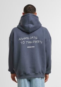 Dark blue oversized hoodie made of cotton. Features a large graphic text "ALWAYS LATE TO THE PARTY" and brand name "ANOTHER COTTON" on the back.