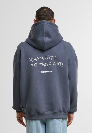 Haut de survêtement oversized bleu foncé en coton. Dispose d'un grand texte graphique "TOUJOURS EN RETARD À LA FÊTE" et du nom de la marque "ANOTHER COTTON" au dos.