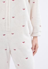 Monos de felpa blanca con bordado de lazo rojo, que presentan un cierre frontal y mangas largas. Textura suave y corte holgado.
