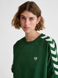 Jeune femme aux cheveux courts et ondulés portant une chemise vert foncé avec des rayures blanches en chevron sur les manches et un petit logo d’abeille sur la poitrine.