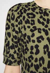 Top de color verde oliva con una tela texturizada que presenta un estampado de leopardo negro. Tiene mangas cortas y un ligero detalle abullonado en los hombros.