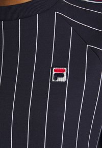 Camiseta deportiva azul marino con rayas verticales blancas y cuello acanalado; presenta un pequeño logo bordado en rojo, blanco y azul.