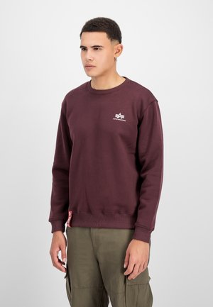 Ung mand iført en rødbrun Alpha Industries sweatshirt og olivengrønne cargobukser, stående mod en ensfarvet lys baggrund.