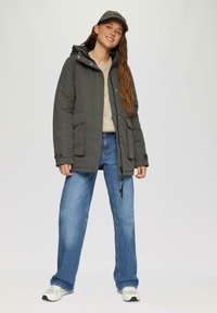 Grijze gewatteerde jas met een capuchon, voorzakken en rits. Gekaapt over een lichte trui en blauwe wijde jeans. Vergezeld door een grijzepet.