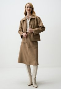 Veste courte beige avec un col cranté et des poches avant, assortie à une jupe longueur genoux ; le modèle porte des bottes beiges montantes.