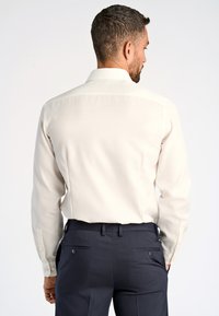 Camicia lunga a maniche beige chiaro con colletto standard, tessuto liscio, cuciture dettagliate e polsini con bottone, abbinata a pantaloni eleganti blu navy.