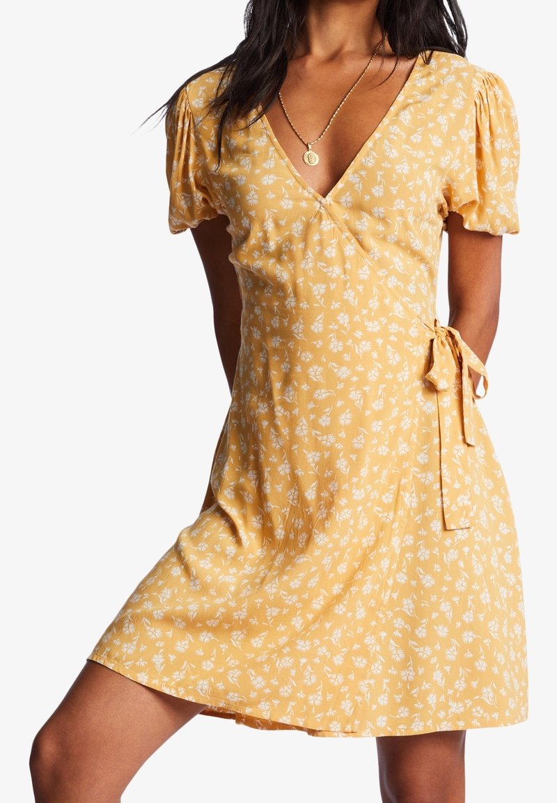 Robe portefeuille jaune à motifs floraux, manches courtes bouffantes, motifs de fleurs blanches, avec un détail de nouage sur le côté. Tissu léger avec une coupe décontractée.