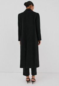 Pitkä musta räätälöity takki, jossa on jämäkkä kaulus, sileä pinta ja pystysaumat, yhdistettynä mustiin housuihin ja korkeakorkoisiin kenkiin.