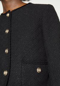 Černá texturovaná bunda s zlatými knoflíky, kulatým výstřihem a přední kapsou s dekorativním prošitím. Syntetický materiál s leskem.