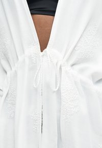 Κοντινή λήψη λευκού υφάσματος με υφή και κεντήματα με λουλούδια, δεμένο στη μέση, αποκαλύπτοντας καφέ δέρμα και μαύρα ρούχα από κάτω.