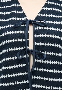 Κοντινό πλάνο υφάσματος πλεκτού με ζιγκ-ζαγκ σε ναυτικό μπλε και λευκό, με δύο δεμένες φιόγκες από ναυτικό μπλε κορδέλα κατά μήκος του κεντρικού ανοίγματος ενός ενδύματος.