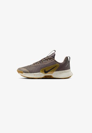Brown athletic shoe with mesh upper, yellow accents, textured sole, and visible speckles. Lace-up design with a pull tab on the heel.