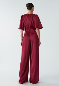 Mujer con el cabello recogido en un moño, vistiendo un mono de satén burdeos con piernas anchas y mangas acampanadas hasta los codos, de pie frente a un fondo liso.