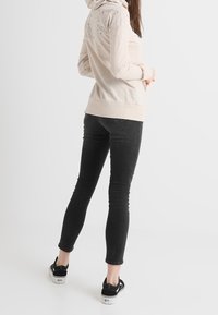 Beige hoodie med en strukturerad yta och flerfärgade mönsterade inslag på baksidan, tillsammans med slimma svarta jeans och svarta skor.