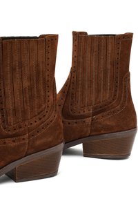 Brązowe zamszowe botki za kostkę z pionowym żeberkowaniem, ozdobnymi perforacjami i słupkowym obcasem. Posiadają zaokrąglony czubek i kontrastowe przeszycia.