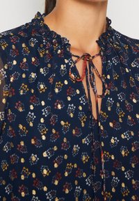 Marinblomstrad blus med sammetskrage, prydd med flerfärgade blommor och gyllene detaljer. Knytning i halsen, genomskinliga tygpaneler vid ärmarna.