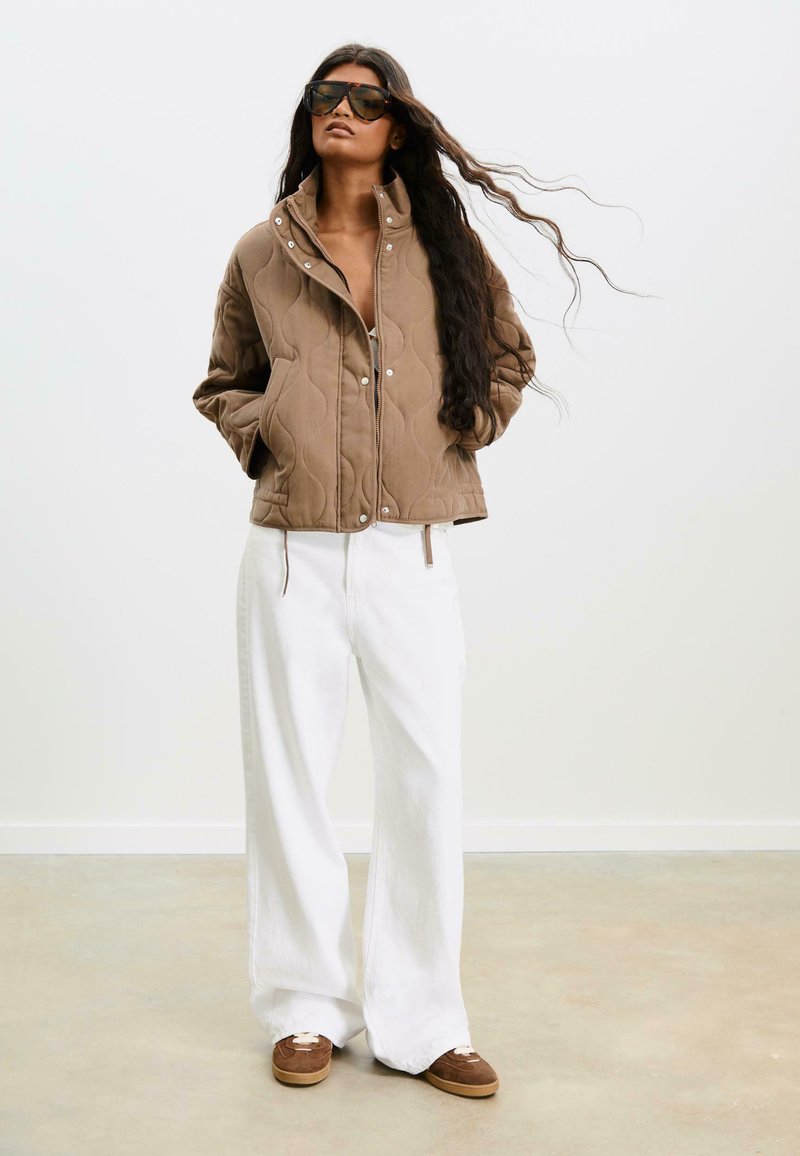 Femme avec de longs cheveux portant une grande veste matelassée marron, un pantalon large blanc, des baskets marron et de grandes lunettes de soleil sombres, se tenant à l'intérieur.